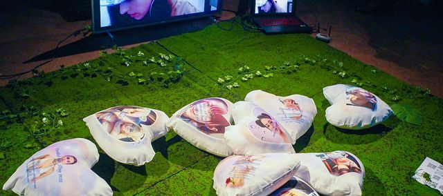 installation image of the work, several cushions with girl images printed on them are laying on a piece of artificial grass.
